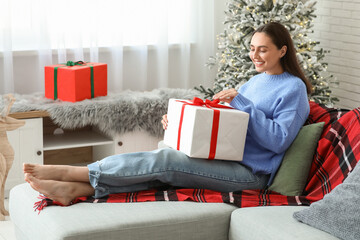 Sticker - Beautiful young woman with Christmas gift box sitting on sofa in living room