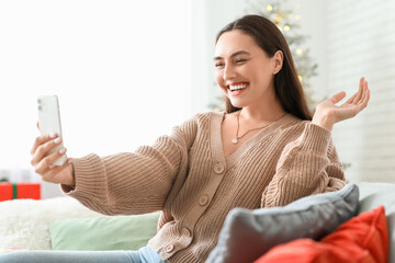 Sticker - Beautiful young woman taking selfie and sitting on sofa in living room. Christmas celebration