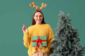 Poster - Young pretty woman in reindeer horns with Christmas tree and gift box on green background