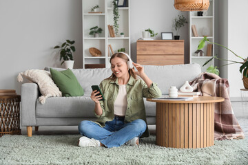 Sticker - Young woman in headphones with mobile phone sitting on carpet at home