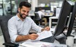 © Mavita - A smiling man working at his desk, reviewing documents with a computer in a modern office setting.