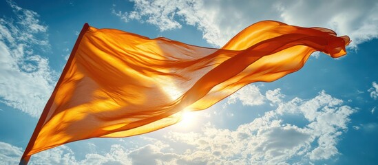 Wall Mural - A vibrant orange flag billows in the wind against a bright blue sky with fluffy white clouds. The sun shines brightly through the fabric, creating a warm and inviting glow.