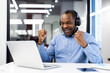 © Liubomir - Happy man wearing headphones cheers while watching sports match on laptop in modern office. Displays excitement and success, representing joy and professional achievement.