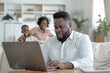 © Руслан Галиуллин - remote work at home, black man is sitting working at laptop, businessman, family is in the background, freelancer
