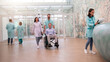 © My Ocean studio - A healthcare team escorts a male patient in a wheelchair through a bright, modern medical facility. Other staff members walk through the clinic, while one reads notes at the reception desk.