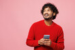 © ViDi Studio - Young smiling happy fun Indian man he wear red sweater casual clothes hold in hand use mobile cell phone look aside isolated on plain pastel light pink background studio portrait. Lifestyle concept.