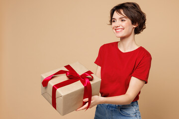 Wall Mural - Side view young happy woman she wear red t-shirt casual clothes hold in hand give present box with gift ribbon bow isolated on plain pastel light beige background studio portrait. Lifestyle concept.