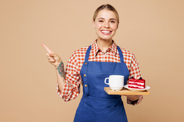 Poster - Young woman barista bartender barman wear blue apron red shirt work in coffee shop hold cake dessert order, cup of tea point aside isolated on plain beige background. Small business startup concept.