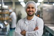© Vadym - Man in sterile uniform stands with arms crossed, smiling at the camera. Hair net worn, indicating a food processing environment. Industrial setting with machinery in the background.