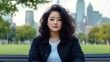 © elimicel - A woman with curly hair sits on a bench in a park, city skyline in the background.