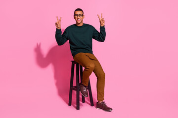 Sticker - Full length photo of handsome good mood guy wear sweatshirt sitting bar chair showing two v-signs emtpy space isolated pink color background