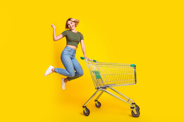 Sticker - Full size photo of overjoyed girl dressed khaki crop top hold shopping cart jump raising fist up isolated on yellow color background