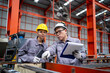 © KANGWANS - Two workers in orange and grey clothing are looking at a piece of machinery. One of them is holding a clipboard