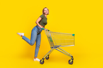 Poster - Full size photo of positive girl dressed khaki crop top jeans walk to empty space with shopping cart isolated on yellow color background