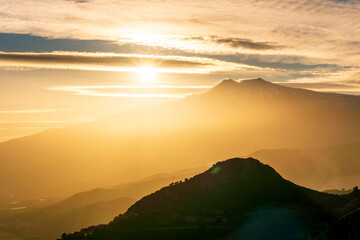 mysterious landscape of great erupting volcano with smoke from craters , clouds of gas and snow on slopes in orange light of sunset. eruption of vulcan