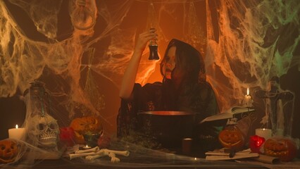 A witch in black clothes is cooking a potion from a spell book in a scary kitchen with pumpkins and a human skull and bones.