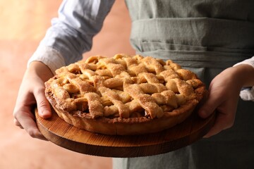 Sticker - Woman holding wooden board with tasty homemade apple pie on light coral background, closeup