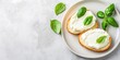 © MdBaki - On a grey background, a platter containing cream cheese, bread pieces, and basil leaves is arranged.