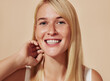 © Artem Varnitsin - Young happy female with blond hair looking at camera in a studio. Beautiful smiling woman with freckles adjusting her hair over a beige backdrop.