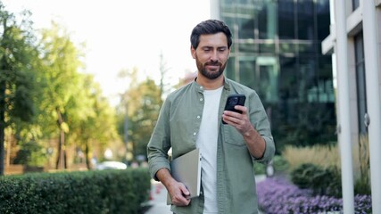 A young businessman is walking outside an office carrying a laptop and smiling at his phone. He's dressed casually and the urban scene is contemporary and bright.