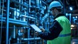 © Anna - A safety-conscious worker wearing a hard hat and high-visibility vest examines information on a tablet while surrounded by industrial machinery in a low-light environment