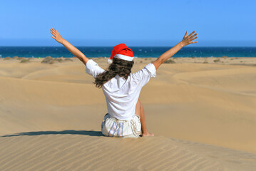 A girl in a dune wearing a Santa hat with hands up. Back view. The concept of a Christmas vacation in hot countries, at the sea. Dune near the sea in Maspalomas, Gran Canaria