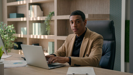 Portrait of serious African American man entrepreneur commercial business work at office male businessman CEO chief working with laptop computer typing data remote job looking at camera confident face