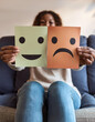 © anandart - Selective focus of female hands holding happy and angry face paper sit on sofa. Feedback rating, emotional intelligence, balance emotion control, mental health assessment, bipolar disorder concept
