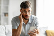 © Home-stock - Shocked indian guy looking at cellphone screen, sitting on couch at home, holding phone and touching his head, reading bad news or message, copy space
