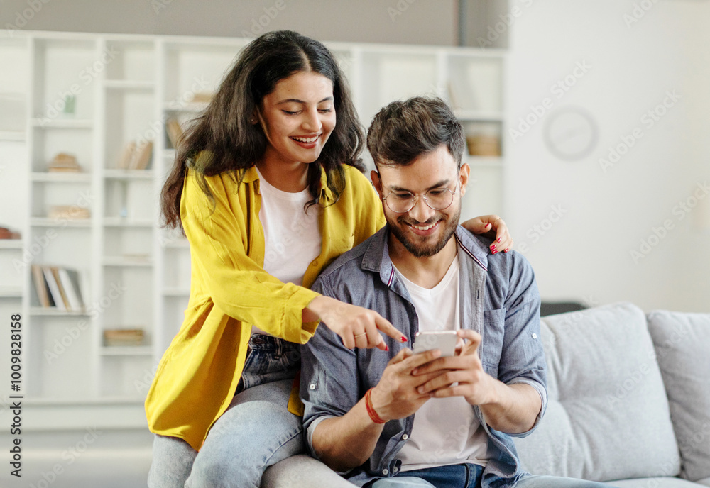 Happy young indian couple using smartphone, wife pointing at cellphone ...