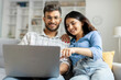 © Home-stock - Happy indian couple using laptop, surfing web, woman pointing at screen, sitting on sofa.