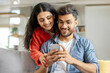 © Home-stock - Cheerful indian couple using cellphone, shopping online, spouses sitting on sofa at home interior