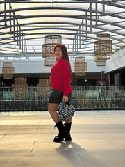 Millennial woman poses confidently in an open, modern environment, wearing a vibrant red sweater and black boots. Joyful expression captures a sense of freedom and individuality