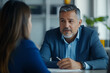 © wolfhound911 - A senior Hispanic bank manager attentively listening to a young client in a modern office setting. The bright space features minimalist decor, fostering an open and professional environment for