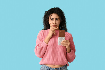 Wall Mural - Thoughtful young African-American woman with sweet chocolate bar on blue background