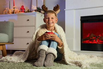 Wall Mural - Little boy in reindeer horns with cup of cocoa sitting near fireplace at home on Christmas eve