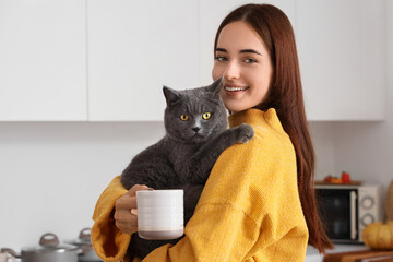 Wall Mural - Young woman with cup of tea and cute British cat in kitchen on autumn day