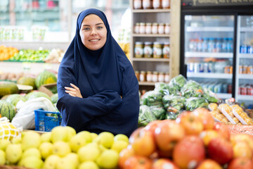 Smiling young muslim female shopper wearing traditional islamic abaya dress and chador standing among produce stands with colorful fresh fruits and vegetables in store