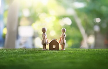 Two wooden figurines stand by a miniature house on a bed of green grass.