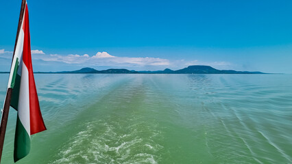 Hungarian flag waving proudly in the wind against backdrop of a calm Balaton lake in Balatonmariafurdo, Hungary, Europe. Tranquil water create a peaceful and patriotic atmosphere. Boat ferry tour