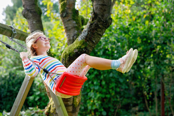 Happy beautiful little school girl having fun on swing in domestic garden. Cute healthy child with eyeglasses swinging under green trees on sunny day. Kidlaughing and crying.