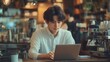 © Aziz - A young man in a white shirt is sitting at a table in a coffee shop working on his laptop.