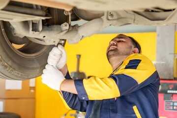 Mechanic working under a car in automotive workshop, wearing a yellow uniform and gloves, focused on repairing vehicle, showcasing professional expertise and attention to detail in maintenance work.