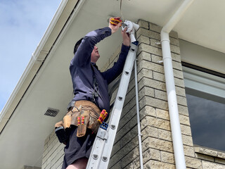 Caucasian man wearing a leather tool belt, holding pliers while standing on a ladder installing a security sensor light to the underside of a soffit on the corner of a brick clad house in New Zealand