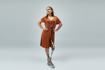 A confident woman stands with her hands on her hips, wearing a brown dress and a prosthetic leg.