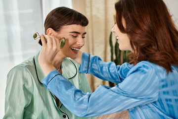 A young lesbian couple enjoys a relaxing morning together, one using a jade roller on the other face.