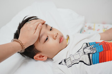 A young boy lies in bed while a hand checks his forehead for fever, showcasing care and concern in a nurturing environment.