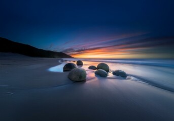 Aufkleber - A group of smooth, round boulders rest on the beach at sunset, with the ocean receding in the background.