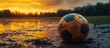 © GoDress - A muddy soccer ball sits in a puddle of water at sunset.