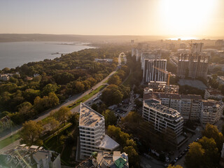 Aerial view of a seaside city with modern buildings, lush greenery, and the calm blue sea in the background. The golden hour light enhances the peaceful urban and coastal landscape